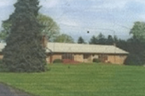 A ranch-style house with a light-colored roof sits amidst a large, lush green lawn. A tall evergreen tree is on the left, under a partly cloudy sky.