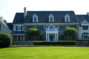 Large suburban house with stone facade, symmetrical windows, and white columns. Manicured lawn, flower beds, and trimmed hedges create a serene feel.