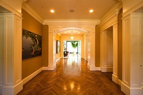 Elegant hallway with warm lighting, parquet flooring, and white columns. A painting hangs on the left wall. An archway leads to a bright room.