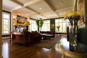 A spacious, warmly lit living room with large windows, a brown sofa, a coffee table, framed photos, and flower vases, creating a cozy, inviting atmosphere.