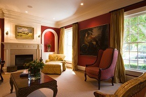 Elegant living room features deep red walls and golden drapes. A cozy fireplace and classic armchairs add warmth, while large art pieces provide sophistication.