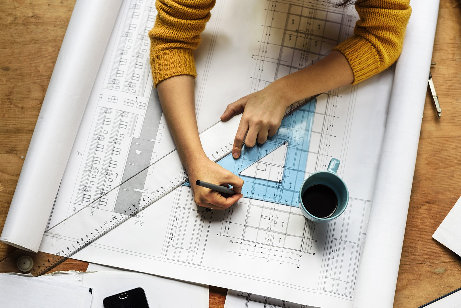 A person draws architectural plans on a desk using a pencil and triangle ruler. They're wearing a yellow sweater. A blue mug and smartphone are nearby.