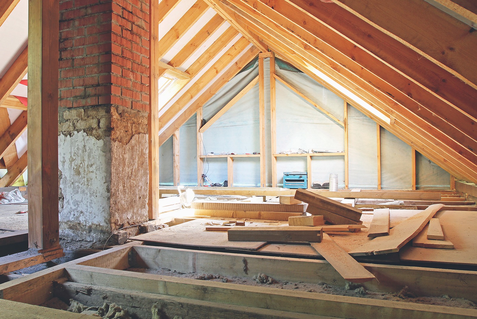 Sunlit attic under renovation with wooden beams and scattered planks. A red brick column stands to the left, emitting a warm, rustic atmosphere.
